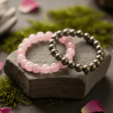 Two beaded bracelets, one pink and one silver, on a stone surface with greenery and petals.